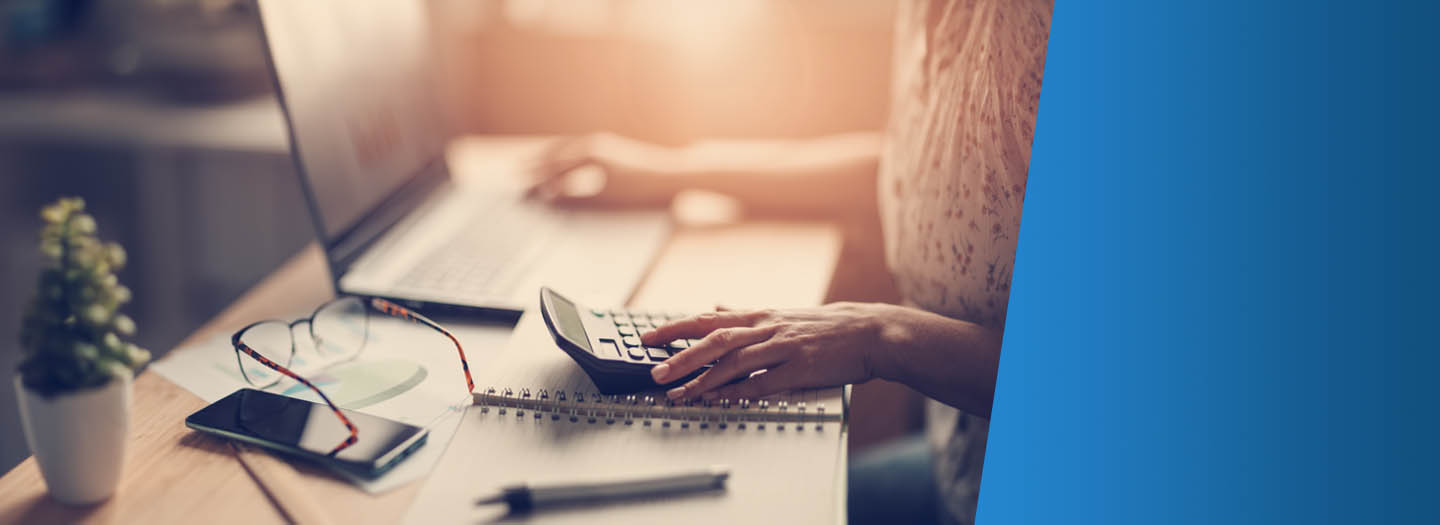 Desk covered with a laptop, calculator and papers, woman's hand preparing to utilize calculator